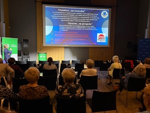 Sala, na krzesełkach, tyłem do zdjęcia siedzi publiczność. Na wprost nich na ścianie jest rzutnik, wyświetlana jest prezentacja. Pod ekranem stoi policjantka, mówi do mikrofonu.