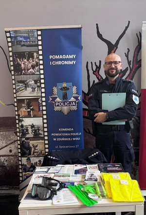 Policjant pozuje do zdjęcia przy stoisku informacyjnym. na stoliku leżą ulotki, odblaski.