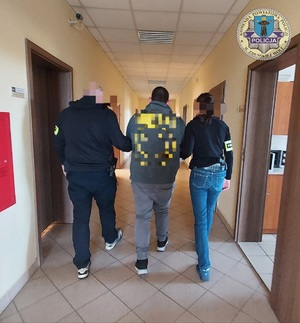 Korytarz w komendzie. Po lewej stronie policjant, a po prawej policjantka, między nimi zatrzymany. Policjanci prowadzą go tyłem do zdjęcia.