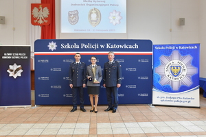 Dwóch policjantów i policjantka pozują do zdjęcia na tle ścianki z napisem Szkołą Policji w Katowicach. Policjantka trzyma w ręku puchar.