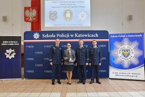 Trzech policjantów i policjantka pozują do zdjęcia na tle ścianki z napisem Szkoła Policji w Katowicach. Policjantka trzyma w ręku puchar.