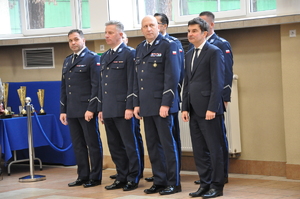 W dwuszeregu stoją umundurowani policjanci i jeden mężczyzna w garniturze. W tle na stole stoją puchary.