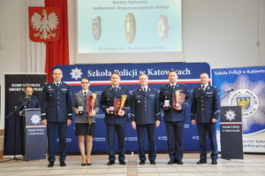 Do zdjęcia pozuje pięciu policjantów i policjantka. Policjantka trzyma w ręku różę i puchar, w tle po lewej stronie stoi za mównicą lektor, mówi do mikrofonu. W tle ściana z napisem Szkoła Policji w Katowicach.