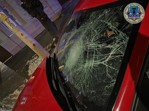 Pęknięta przednia szyba w samochodzie. Noc, w tle chodnik i fragment budynku.