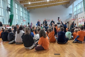 Sala gimnastyczna. Na podłodze siedzą dzieci, podnoszą do góry ręce, zgłaszają się do odpowiedzi, przed nimi stoi troje policjantów.