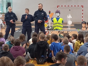 Sala gimnastyczna. Na podłodze siedzą dzieci, podnoszą do góry ręce, zgłaszają się do odpowiedzi, przed nimi stoi troje policjantów.