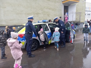Przed budynkiem stoi otwarty radiowóz policyjny, obok stoją policjanci, do samochodu wsiadają dzieci.