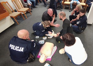 Na podłodze, obok siebie, pochylają się policjanci i cywilne osoby, ćwiczą na fantomach, przyglądają się innym ćwiczącym.