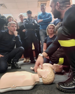 Na podłodze, obok siebie, pochylają się policjanci i cywilne osoby, ćwiczą na fantomach, przyglądają się innym ćwiczącym.