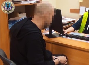 Zatrzymany siedzi przy biurku, ma kajdanki założone na ręce, przed nim siedzi policjant.