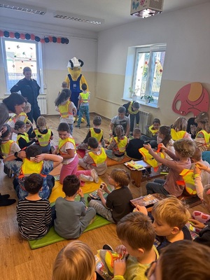 Dzieci w obecności policjantki i maskotki marketu Mrówka zakładające na siebie kamizelki odblaskowe.