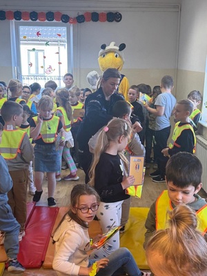 Dzieci z otrzymanymi elementami odblaskowymi oraz policjantka zakładająca dzieciom kamizelki.