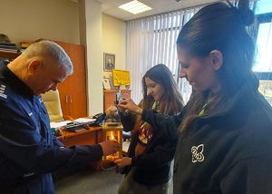 Policjant od palącej świecy podpala drugą świeczkę. Obok policjanta stoją dwie harcerki.