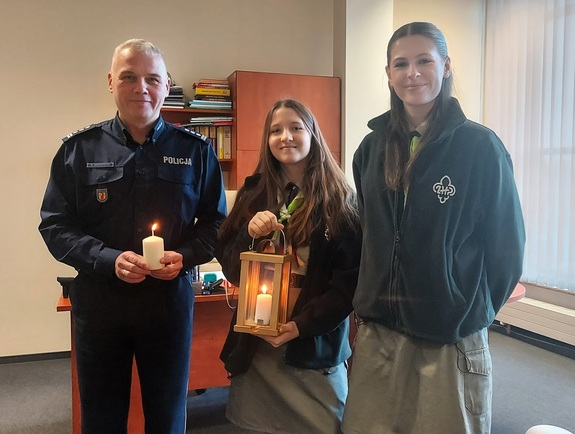 Policjant oraz dwie harcerki pozują z palącymi się świeczkami do zdjęcia.