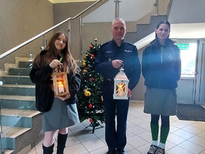 Policjant i dwie harcerki pozują z palącymi się świeczkami do zdjęcia. W tle choinka.