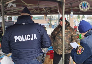 Policjant i strażniczka miejska rozmawiają ze sprzedawcą fajerwerków. Oglądają dokumentację dotyczącą legalności takiej sprzedaży. W tle ustawione do sprzedaży różne petardy i fajerwerki.