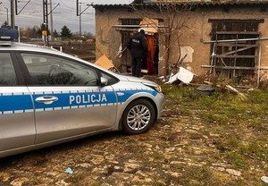 Po lewej stronie zaparkowany radiowóz. W tle opuszczony, zniszczony budynek, policjant zagląda do wnętrza, zamiast drzwi wisi zasłona, okna są zabite deskami.