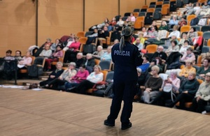 Policjantka stoi na scenie, tyłem do zdjęcia, przodem do publiczności. Na krzesłach siedzą słuchacze.
