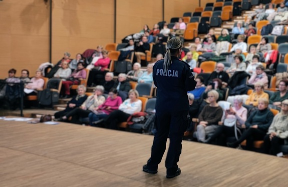 Policjantka stoi na scenie, tyłem do zdjęcia, przodem do publiczności. Na krzesłach siedzą słuchacze.