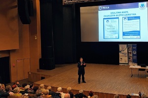 Policjantka stoi na scenie przodem do publiczności, za jej plecami na ekranie wyświetla się zdjęcie dotyczące Krajowej Mapy Zagrożeń Bezpieczeństwa.