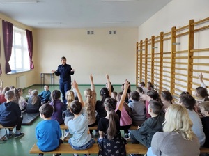 Sala w szkole. Po lewej stronie okna, po prawej drabinki do ćwiczeń. Uczniowie siedzą na ławkach tyłem do zdjęcia, na wprost nich stoi policjantka, mówi do nich. Niektórzy uczniowie podnoszą ręce do góry, zgłaszają się do odpowiedzi.