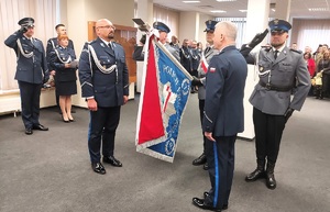 Poczet sztandarowy. Obok sztandaru stoją dwaj komendanci. W tle publiczność.