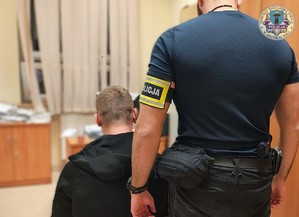 Zatrzymany siedzi na krzesełku tyłem do zdjęcia, koło niego stoi policjant ubrany po cywilnemu, na lewej ręce ma opaskę z napisem Policja.