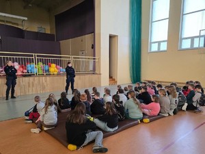 Na sali gimnastycznej, policjantki mówią do siedzących na podłodze przed nimi dzieci.