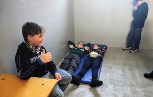 Dwóch chłopców leży na materacu, jeden siedzi na krzesełku przy stoliku. Są w PDOZ.