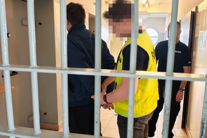 Za kratami stoi zatrzymany, ma z tyłu ręce, policjant zakłada mu kajdanki.