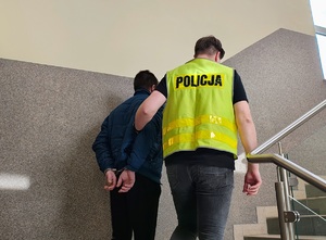 Policjant prowadzi po schodach zatrzymanego, który ma kajdanki na rekach.