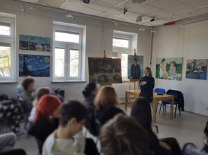 Klasa w szkole, na ścianach wiszą obrazy, stoją na sztalugach. Przed uczniami stoi policjantka, mówi do zebranych.