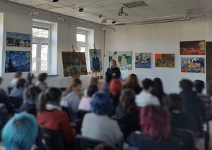 Klasa w szkole, na ścianach wiszą obrazy, stoją na sztalugach. Przed uczniami stoi policjantka, mówi do zebranych.