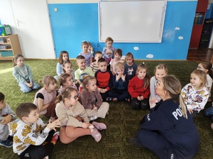 Policjantka siedzi przed dziećmi na krzesełku. Mówi do nich. Dzieci siedzą na podłodze, patrzą na policjantkę, słuchają jej.