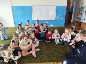 Policjantka siedzi przed dziećmi na krzesełku. Mówi do nich. Dzieci siedzą na podłodze, patrzą na policjantkę, słuchają jej.