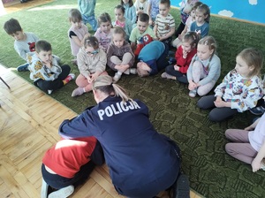 Policjantka jest przed dziećmi. Mówi do nich. Dzieci siedzą na podłodze, patrzą na policjantkę, słuchają jej.