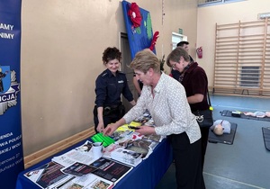 Policjantka przy stoisku profilaktycznym, pokazuje ulotki kobiecie.