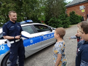 Policjant stoi przy radiowozie, obok niego dzieci, słuchają co do nich mówi.