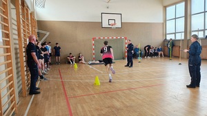 Sala gimnastyczna w szkole. Na sali uczestnicy podczas sportowych zmagań. Są też inni uczestnicy siedzący na ławce i pod ścianą, policjanci sędziują.