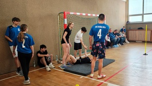 Sala gimnastyczna w szkole. Na sali uczestnicy podczas sportowych zmagań. Są też inni uczestnicy siedzący na ławce i pod ścianą, policjanci sędziują.