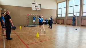 Sala gimnastyczna w szkole. Na sali uczestnicy podczas sportowych zmagań. Są też inni uczestnicy siedzący na ławce i pod ścianą, policjanci sędziują.