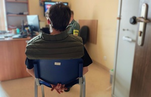 Pomieszczenie, jakiś pokój. Tyłem do zdjęcia na krzesełku siedzi mężczyzna. Ma kajdanki założone na ręce trzymane z tyłu.