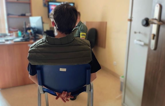 Pomieszczenie, jakiś pokój. Tyłem do zdjęcia na krzesełku siedzi mężczyzna. Ma kajdanki założone na ręce trzymane z tyłu.