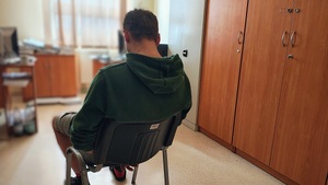 Pomieszczenie, jakiś pokój. Tyłem do zdjęcia na krzesełku siedzi mężczyzna. Ma kajdanki założone na ręce trzymane z tyłu.