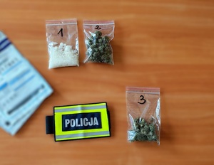 Po lewej stronie dwie koperty na dowody. Po prawo trzy torebki strunowe - jedna z białym proszkiem, a dwie z suszem. Obok nich leży opaska z napisem Policja.
