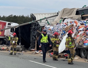 Na drodze idzie policjant, w tle strażacy i rozsypane na drogę, z przyczepy sypkie materiały.