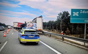 Policyjny radiowóz, w tle uszkodzony pojazd z rozsypanym na drogę towarem.