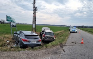 Przy drodze, w rowie, leżą dwa samochody osobowe. W tle widać zaparkowany radiowóz.