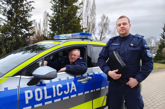 W radiowozie siedzi jeden policjant, wygląda przez okno, a obok stoi drugi, trzyma w ręku aktówkę. Policjanci pozują do zdjęcia.