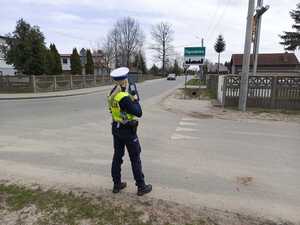 Policjantka stoi na drodze, tyłem do zdjęcia, w rękach trzyma urządzenie do pomiaru prędkości, przed nią tablica z napisem: Ogrodzisko.